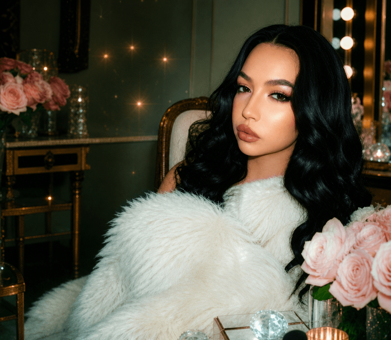 Glamorous woman with long black hair wrapped in white faux fur, amidst pink roses.