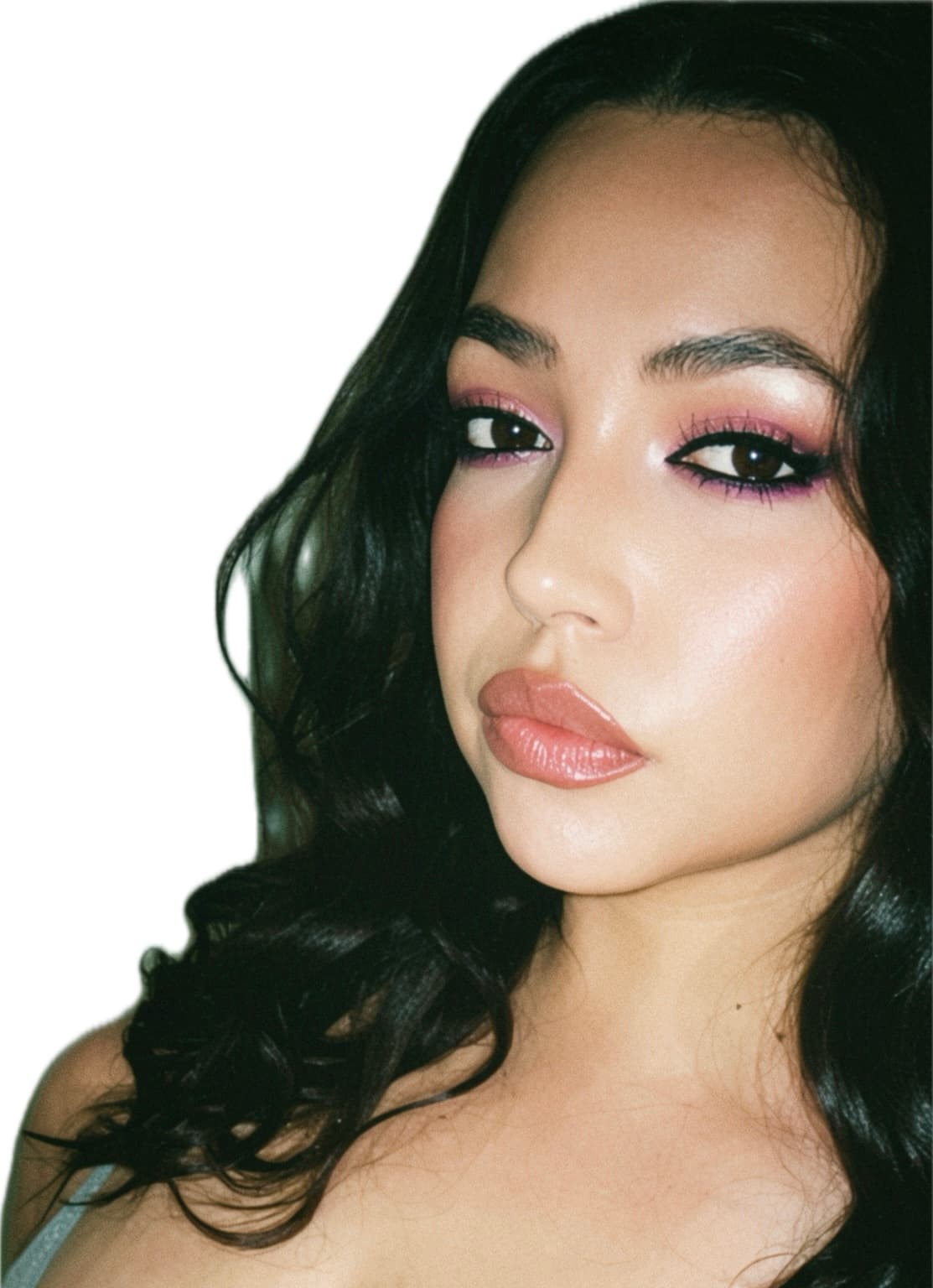 Close-up of a woman with dark wavy hair, pink eyeshadow, winged eyeliner, and glossy lips.